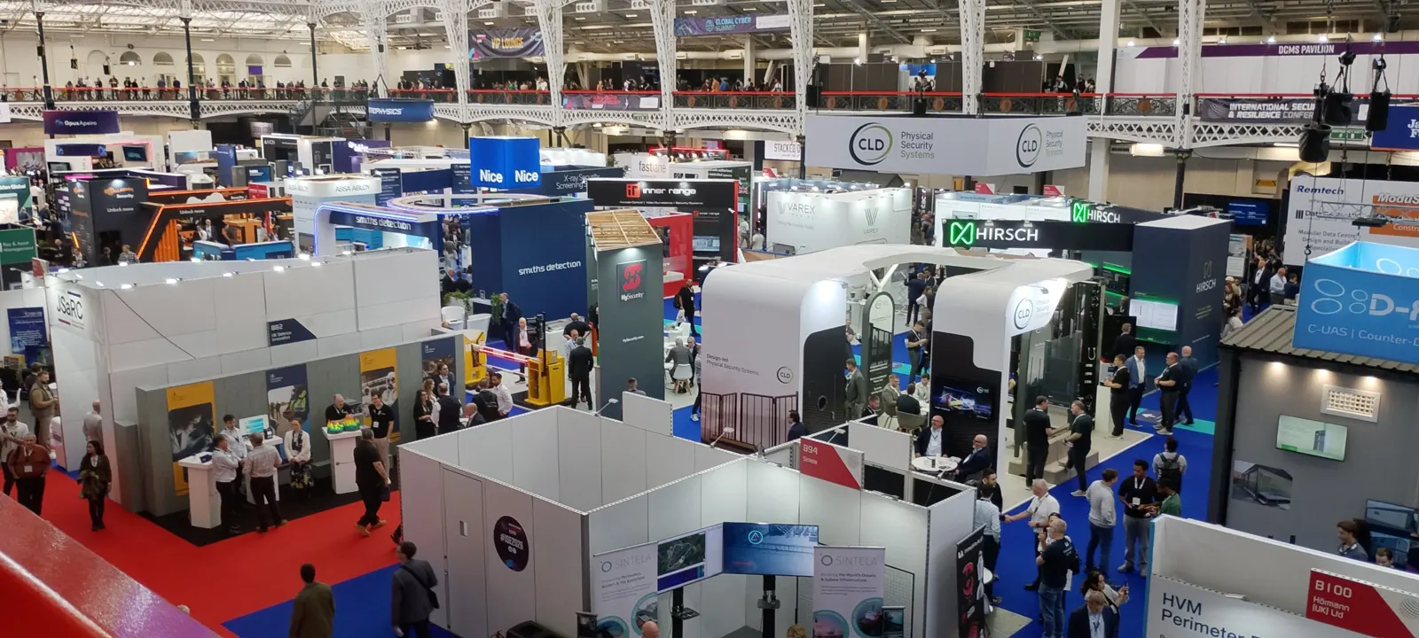 Overhead view of the International Security Expo exhibition floor at Olympia, London, with stands for access control, perimeter protection, screening and counter-drone systems, and the Global Cyber Summit banner on the upper level.