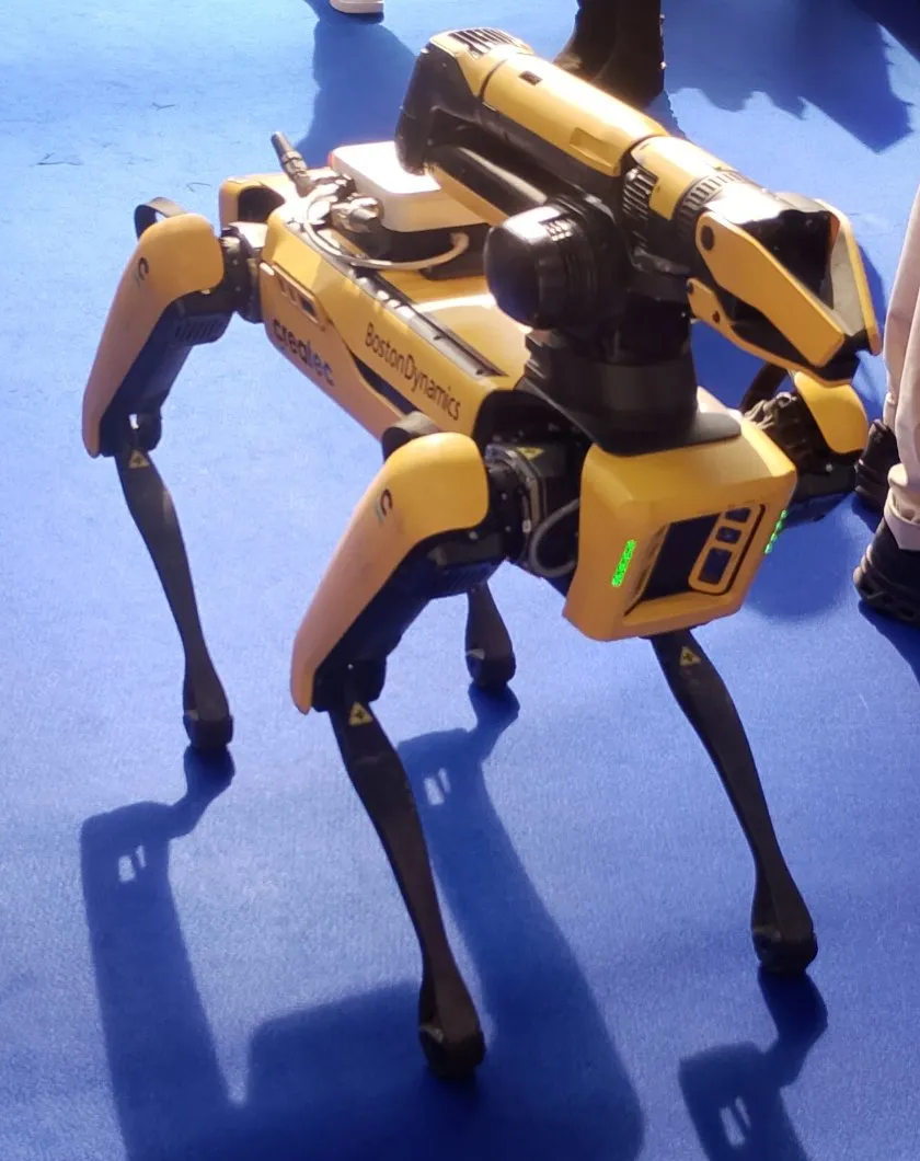 A yellow four-legged Boston Dynamics Spot robot carrying an arm and sensor head, with Createc branding, standing on blue exhibition carpet.
