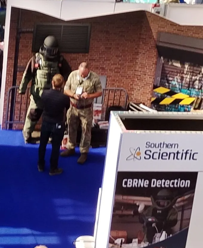 A bomb disposal suit on display against a brick-wall set at a stand in the exhibition hall, with a CBRNe detection banner on the neighbouring stand.